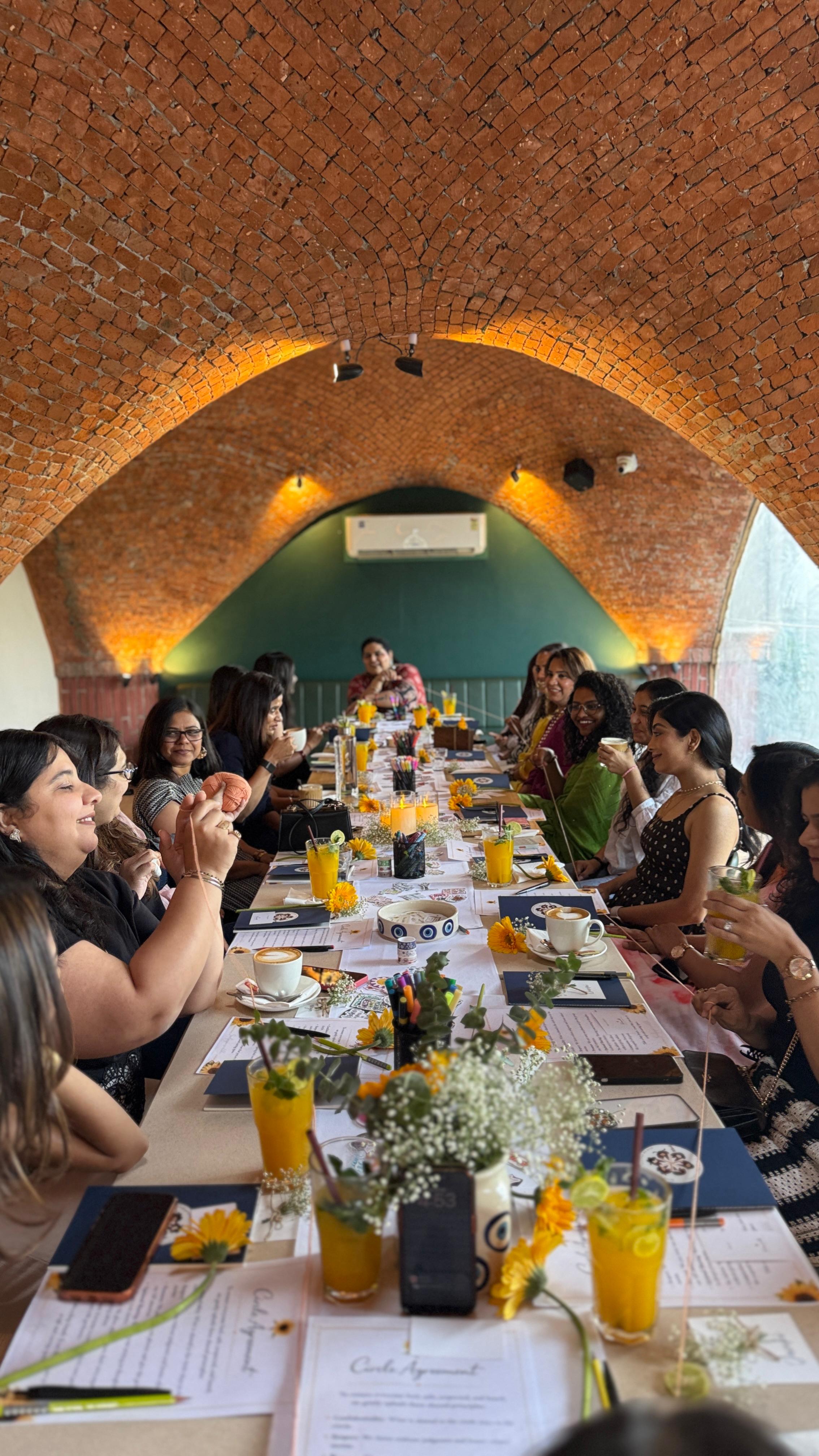 Group of women seated around a long table with drinks and flowers under a brick vaulted ceiling.