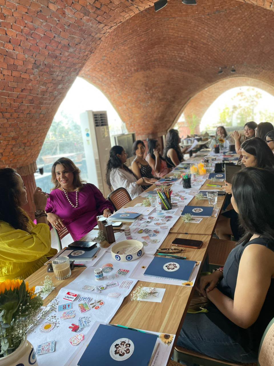 Women seated around a long table under brick arches, engaged in discussion in a bright room.